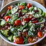 A vibrant Greek Salad Bowl served in a white bowl, topped with crumbled feta and briny olives, perfect for a refreshing summer lunch.