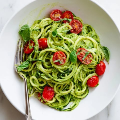 Creamy avocado pesto coats spiralized zucchini noodles, topped with fresh cherry tomatoes and basil for a vibrant, healthy meal.