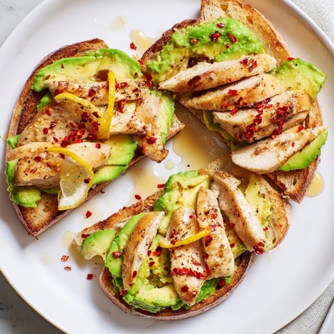 Golden grilled chicken slices arranged over creamy smashed avocado on crispy sourdough toast, topped with chili flakes and honey.